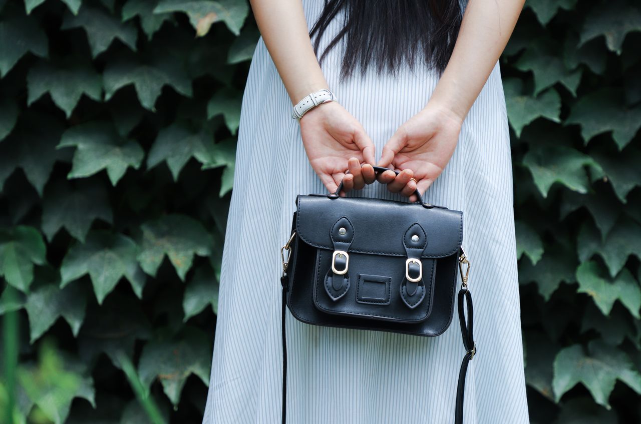 services-02 A woman in a striped dress holds a black leather handbag with lush green leaves in the background.